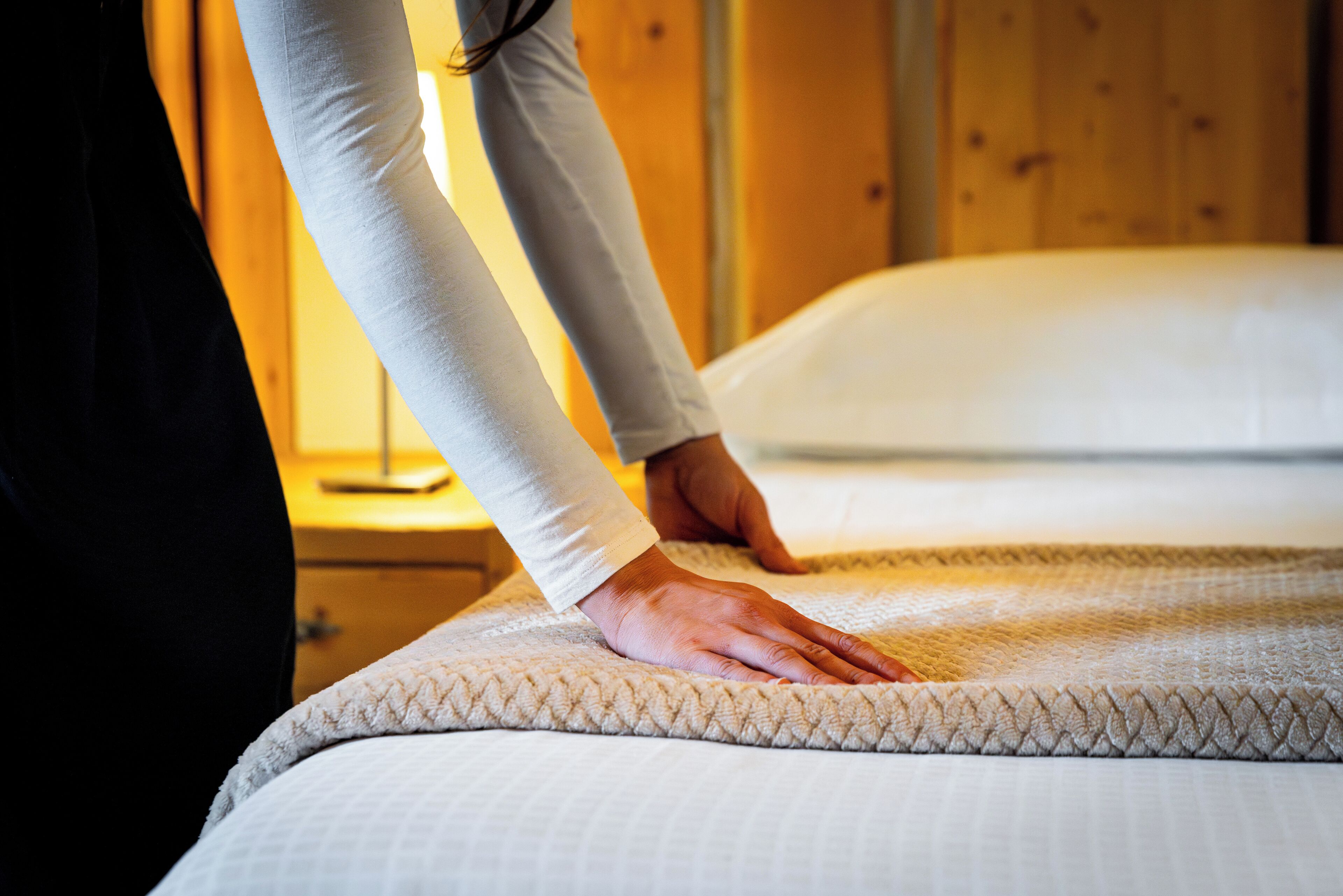 A hand adjusting a bed with a textured white cover in a room with wooden walls.