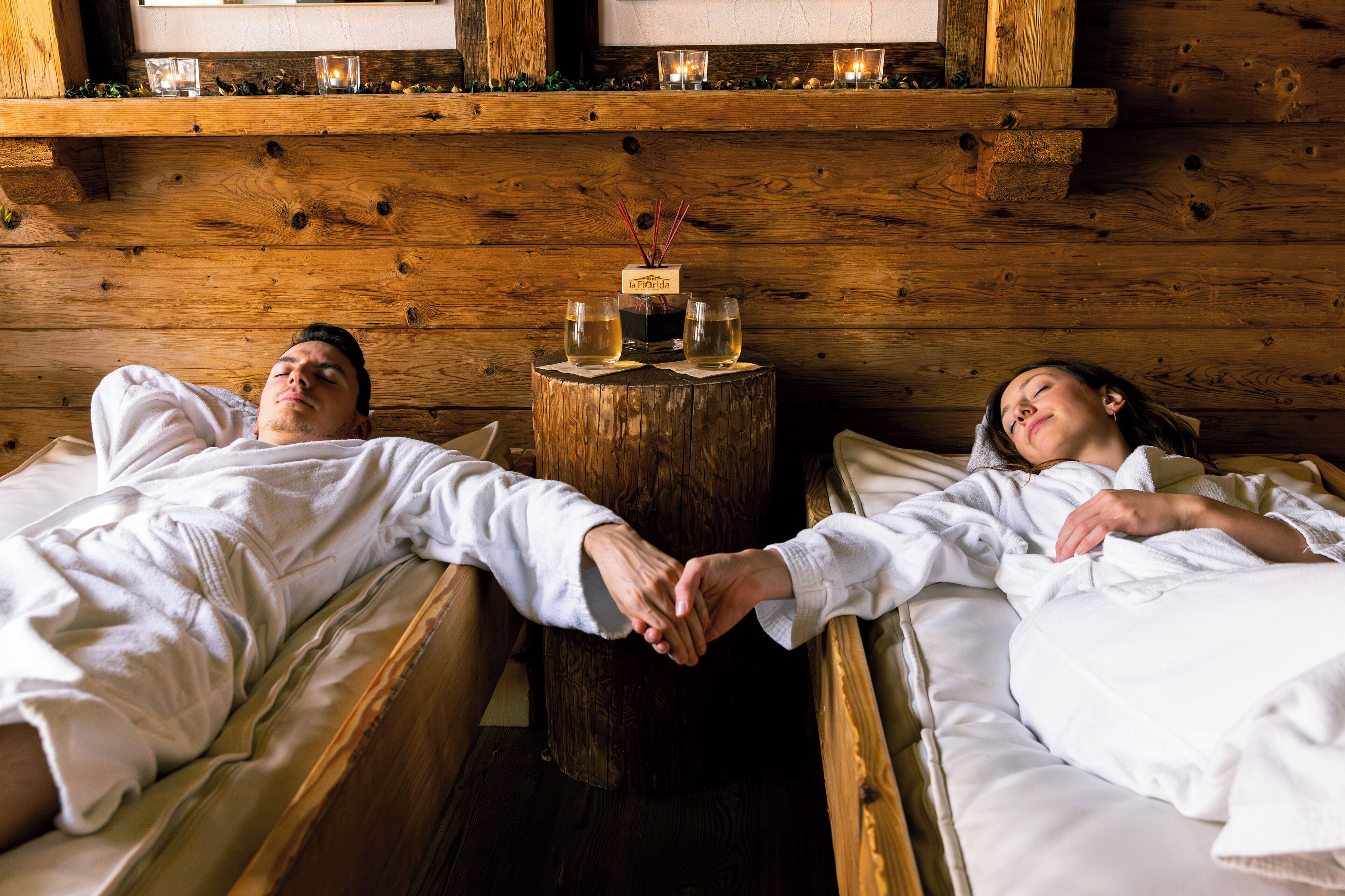 Two people lying in separate beds in a rustic cabin-style room with wooden walls, a small wooden table with two glasses and a candle between them.