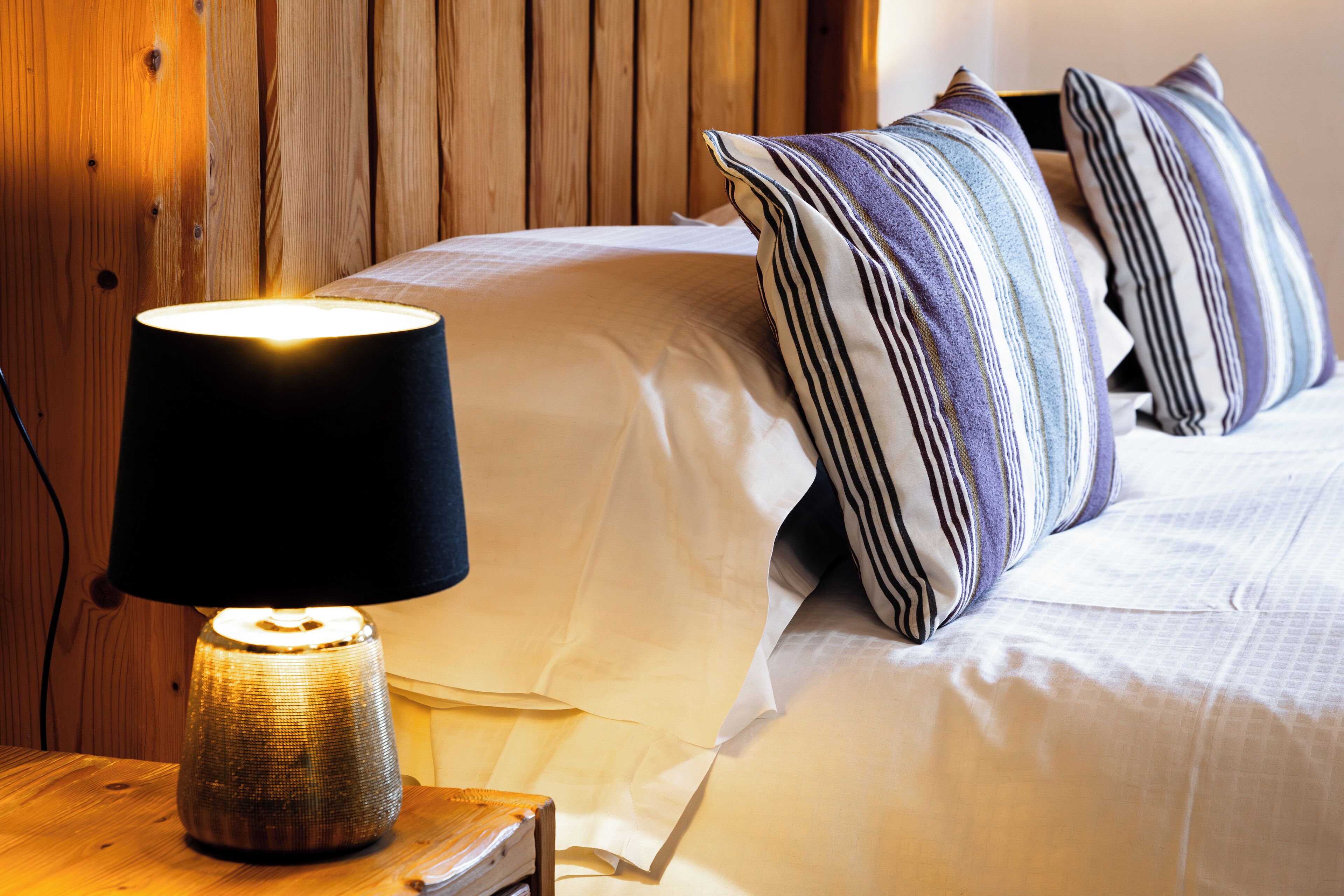 Una cama bien hecha con almohadas a rayas y una lámpara de mesa encendida.