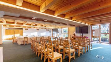 Une salle de conférence dotée de chaises et de tables en bois, de grandes fenêtres et d’un écran de projection.