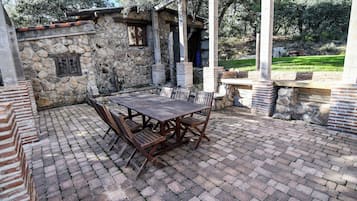 A stone patio with a wooden table and chairs, surrounded by stone walls and a view of trees.