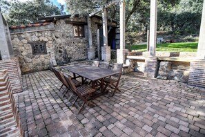 A stone patio with a wooden table and chairs, surrounded by stone walls and a view of trees. - Casa Rural Las Corzas (Casavieja)