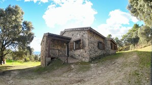 A rustic stone building with a wooden door and windows, situated on a grassy slope with trees in the background.