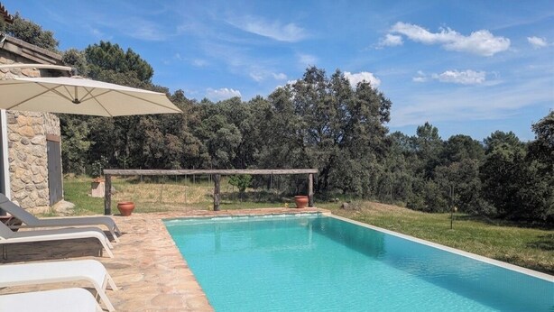 A swimming pool with stone tiles, a pergola with a white umbrella, and a stone building in the background. - Casa Rural Las Corzas (Casavieja)