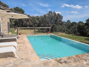 A swimming pool with stone tiles, a pergola with a white umbrella, and a stone building in the background. - Casa Rural Las Corzas (Casavieja)