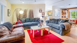 Un salon moderne avec deux canapés, une table basse et un tapis rouge. On aperçoit, au fond, une cuisine avec des meubles en bois et un coin repas.