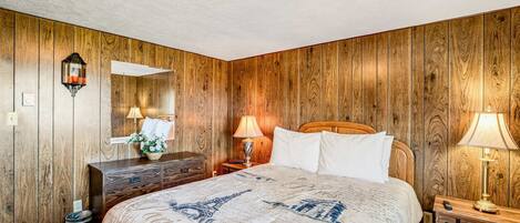 Un dormitorio con un muro revestido de madera, una cama con cubrecama con temática de París, un aparador, una mesita de noche con lámpara y un espejo.