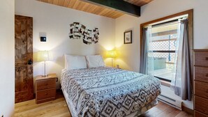 Een slaapkamer met een houten plafond, een bed met beddengoed met een patroon, een nachtkastje met een lamp, een wandthermostaat en een raam met gordijnen.