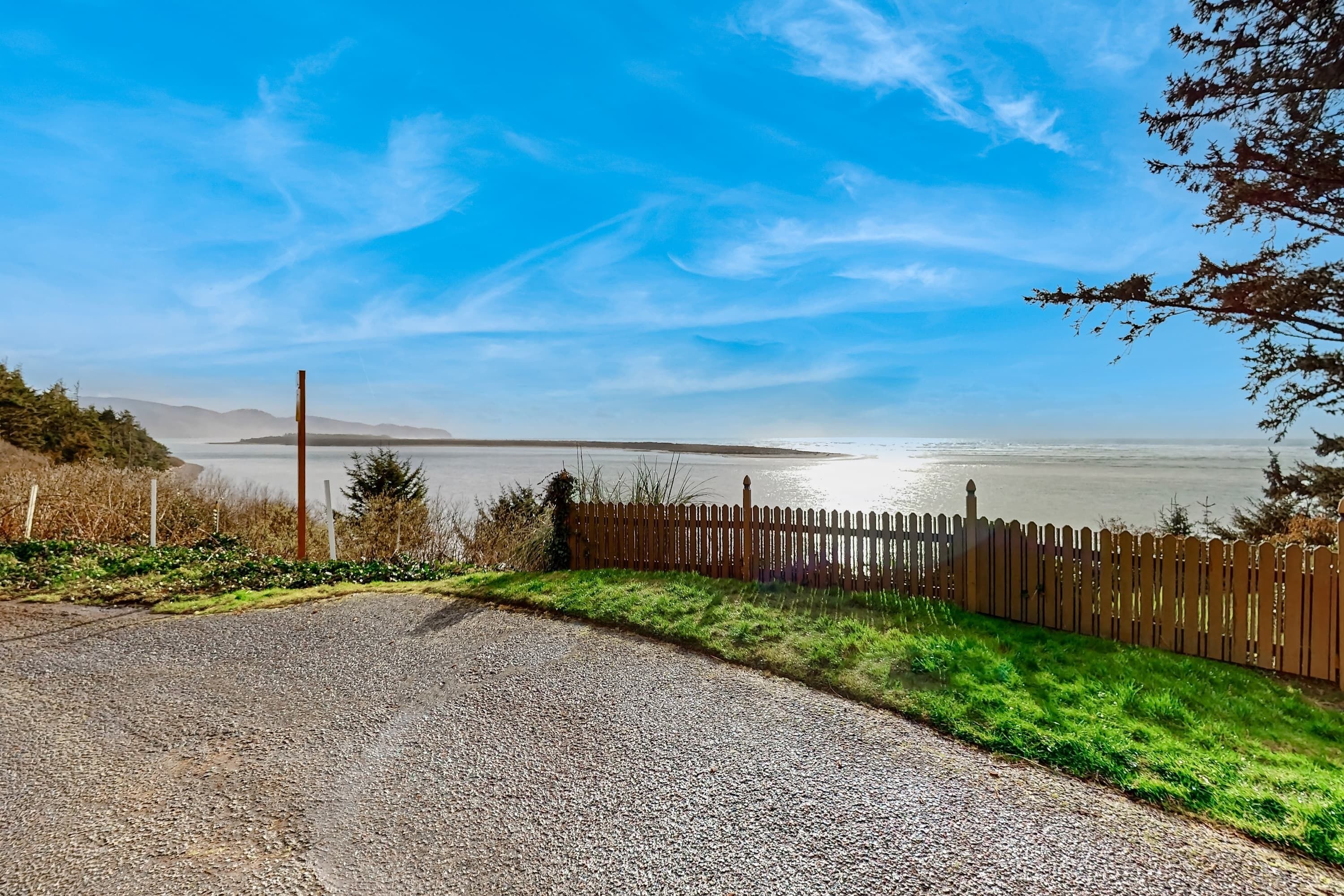 A gravel driveway leading to a wooden fence overlooking a body of water with distant mountains.