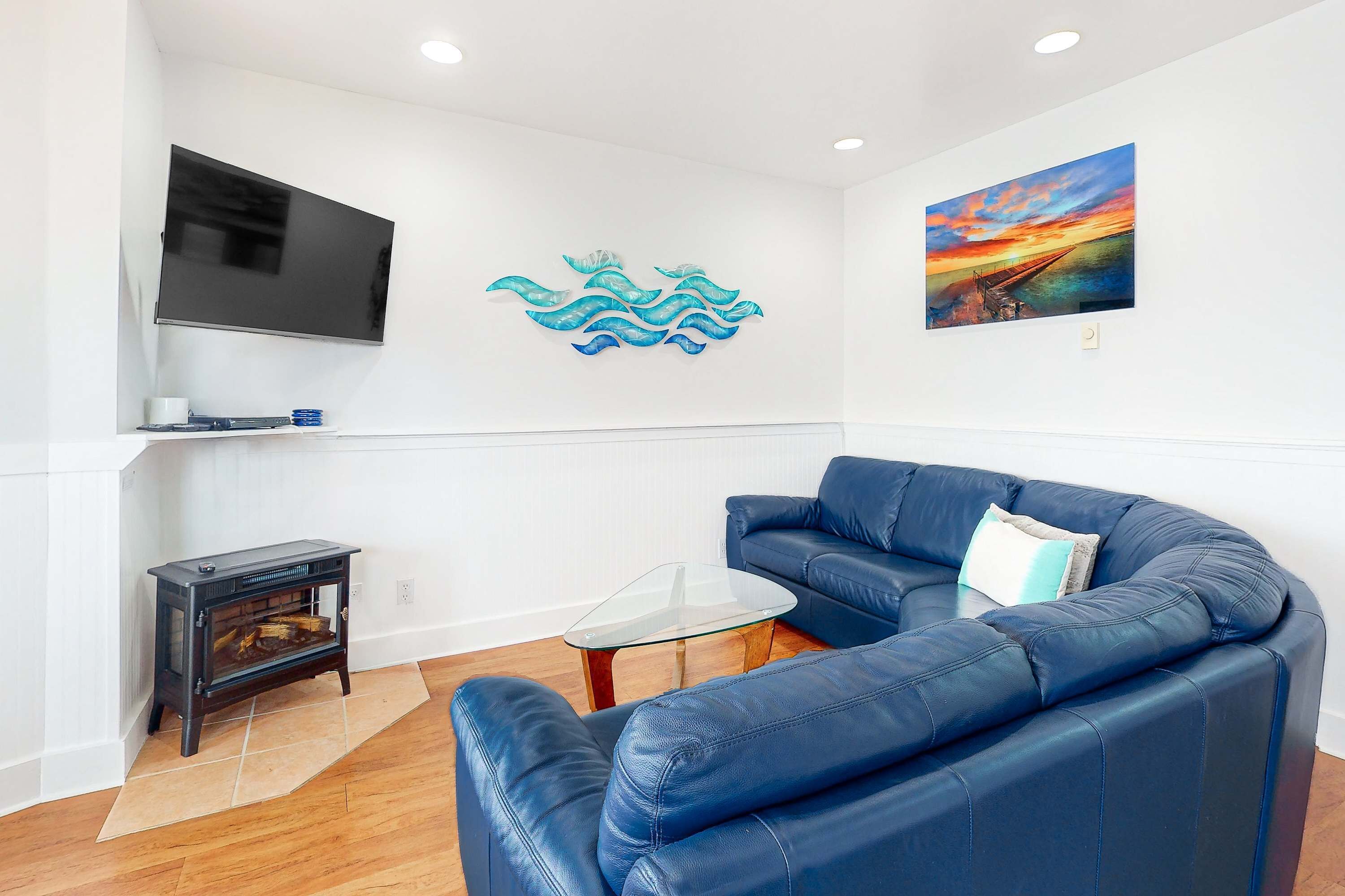 Una sala de estar con un sofá azul, una mesa de centro de vidrio, una chimenea, un televisor y dos obras de arte en la pared.