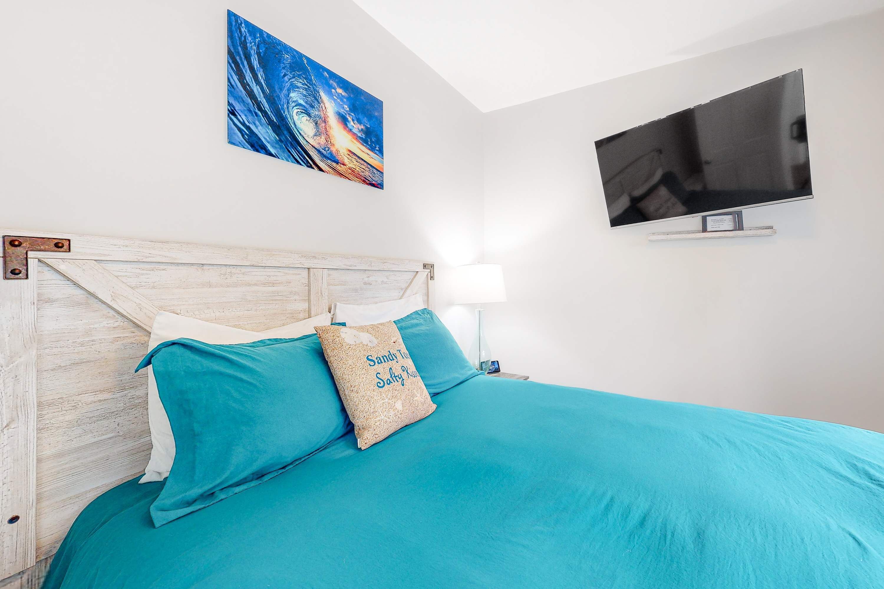 Una cama con ropa de cama color turquesa, cabecera de madera y almohada con texto. Un televisor está montado en la pared sobre la cama.