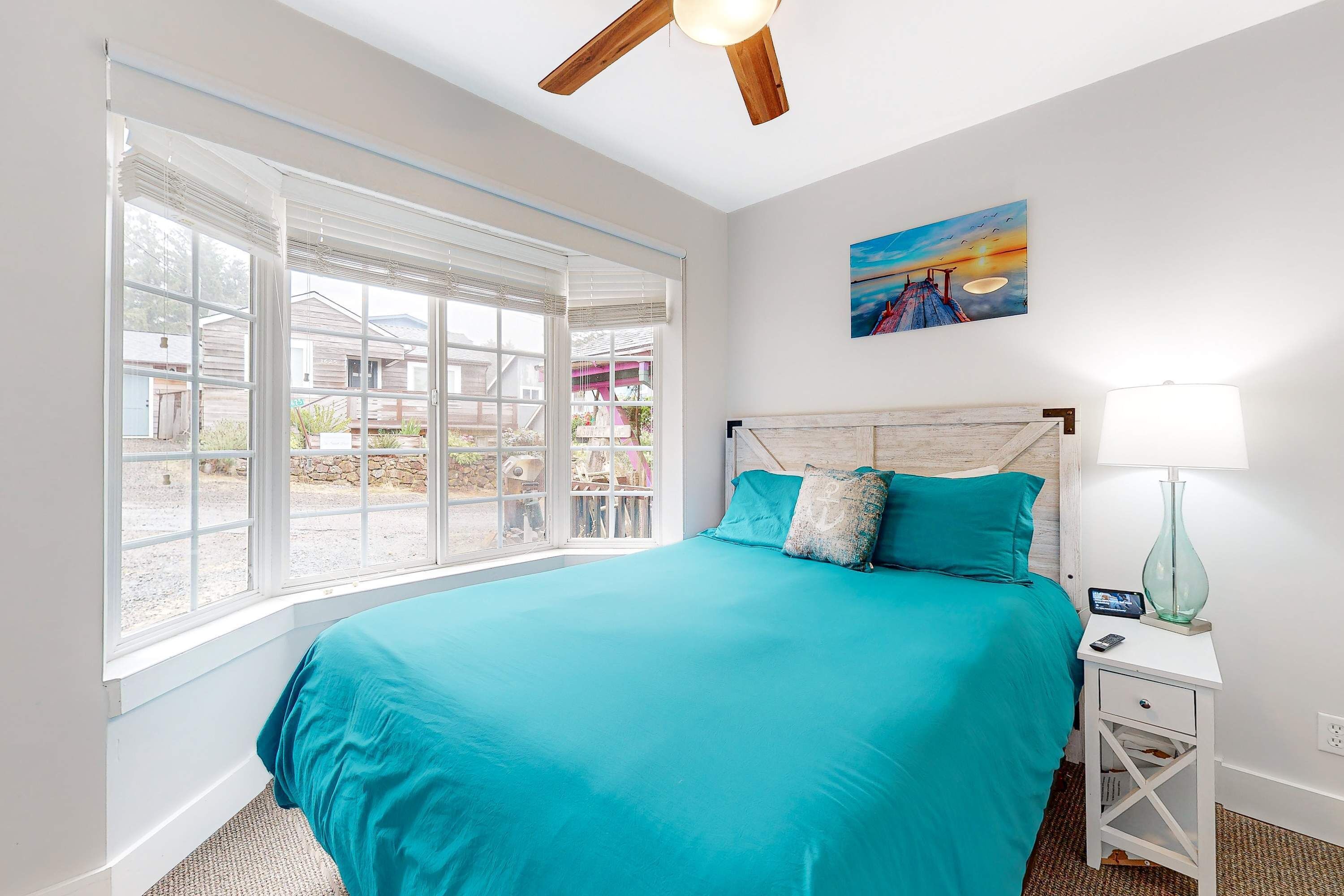 A bedroom with a large bed, a ceiling fan, a nightstand with a lamp, and a painting on the wall.