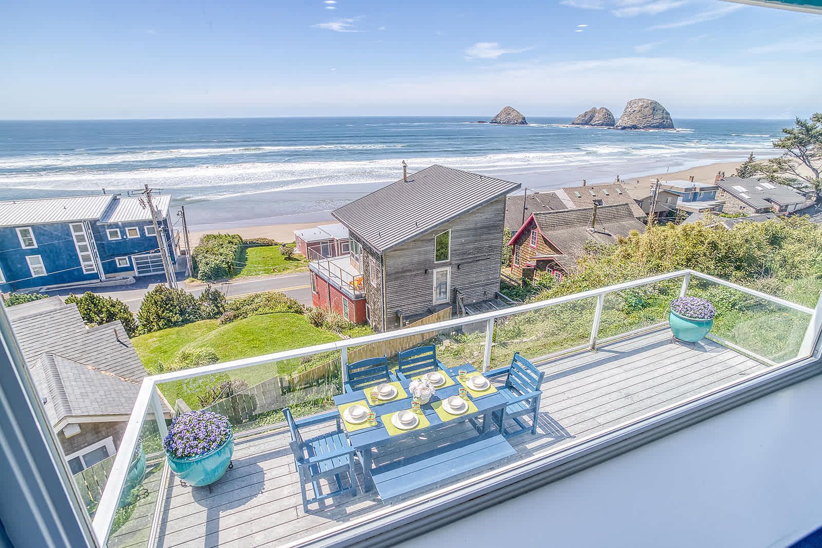 Varanda com mesa posta para refeições, com vista para uma praia com afloramentos rochosos e casas.