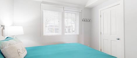 Un dormitorio con colcha color esmeralda, paredes blancas, una puerta blanca y una ventana con persianas.