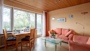 Una sala de estar moderna con techo de madera, ventanas amplias, una mesa de centro de vidrio, un sofá rosa y un comedor con mesa y sillas de madera.