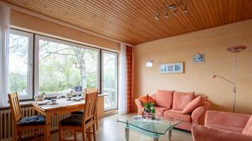 Ein modernes Wohnzimmer mit Holzboden, großen Fenstern, einem Glastisch, einem rosafarbenen Sofa und einem Essbereich mit Holztisch und Stühlen.