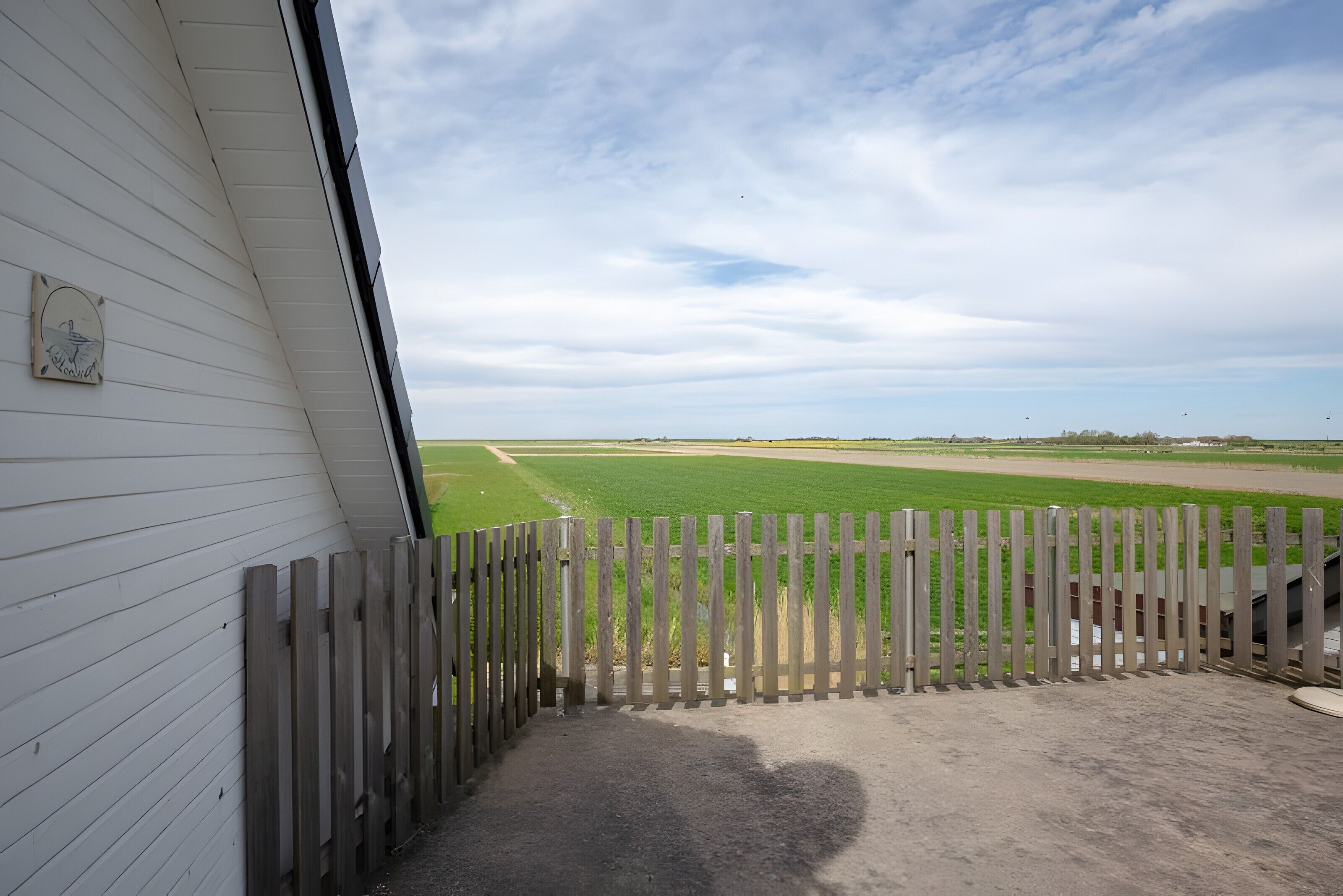 Blick durch einen Holzzaun auf ein grünes Feld, ein weißes Gebäude mit einem Fenster und einem klaren Himmel.