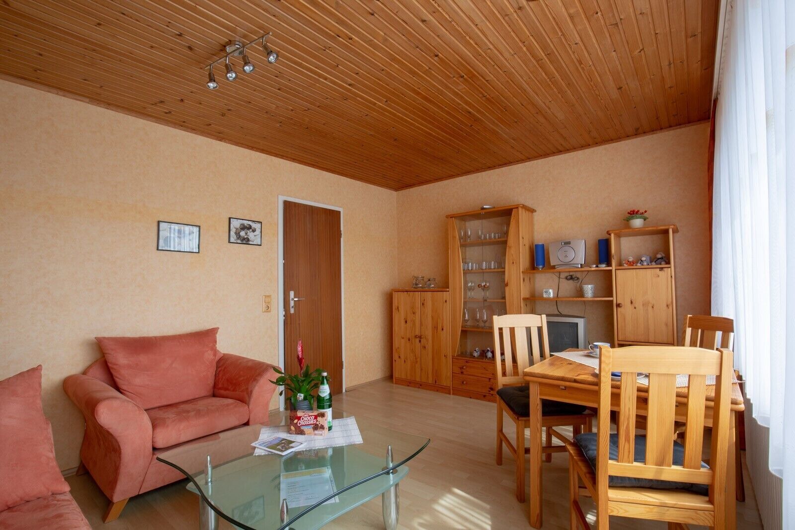 Ein gemütliches Wohnzimmer mit Holzboden, einem Glastisch, einem rosa Sofa, einem Holzesstisch mit Stühlen und einem Holzschrank mit Gläsern.