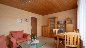 Una sala de estar acogedora con techo de madera, una mesa de centro de vidrio, un sofá rosa, una mesa de comedor de madera con sillas y un aparador de madera con cristalería.