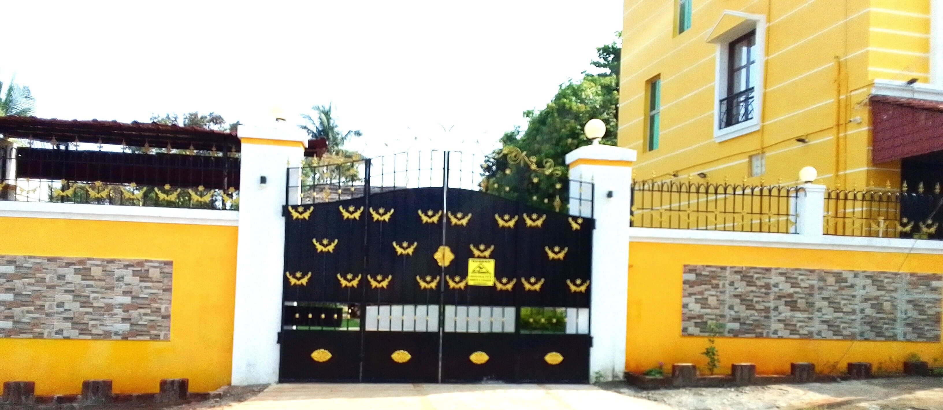 A gated entrance with a decorative gate, a yellow building with a stone facade, and a small balcony with a railing.