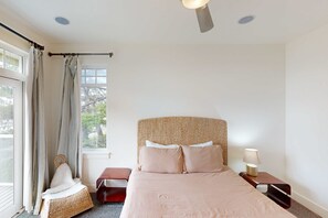Um quarto com cama, mesa de cabeceira, cadeira, janela com cortinas e ventilador de teto.
