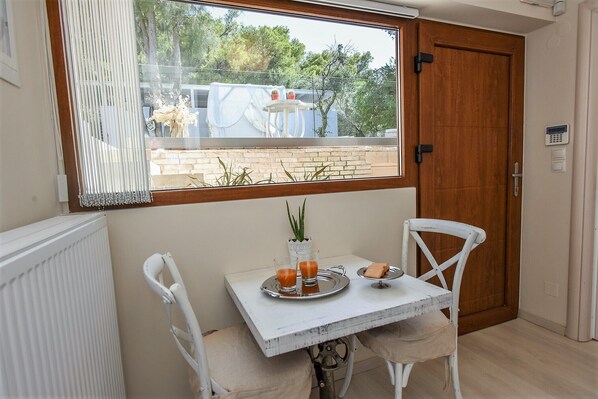 A small dining area with a table and two chairs, a window with blinds, and a view of a building and trees outside. - Villa Evangelia (Chania)