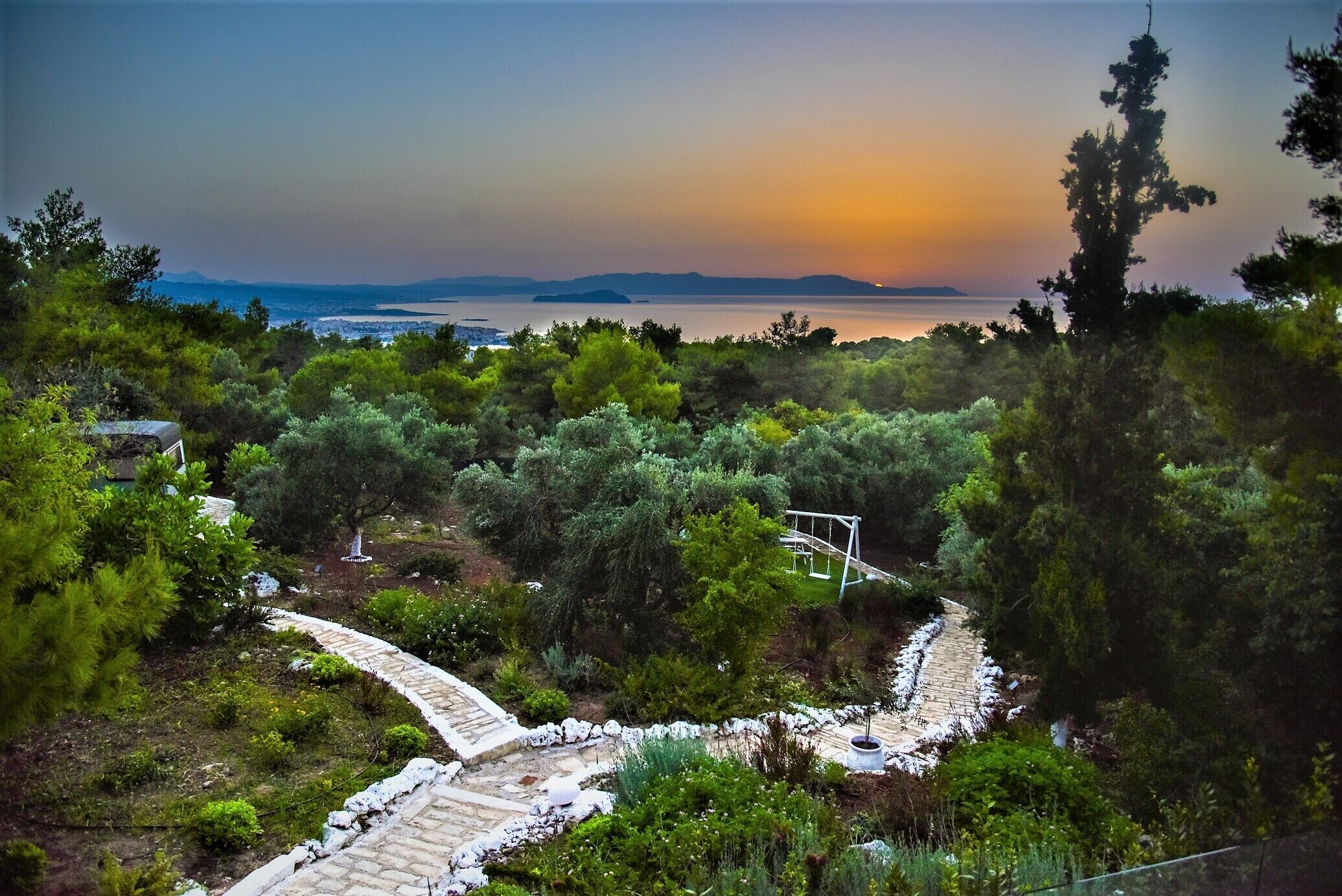 A garden with a stone pathway leading to a gazebo, surrounded by dense greenery and overlooking a body of water with distant landforms.