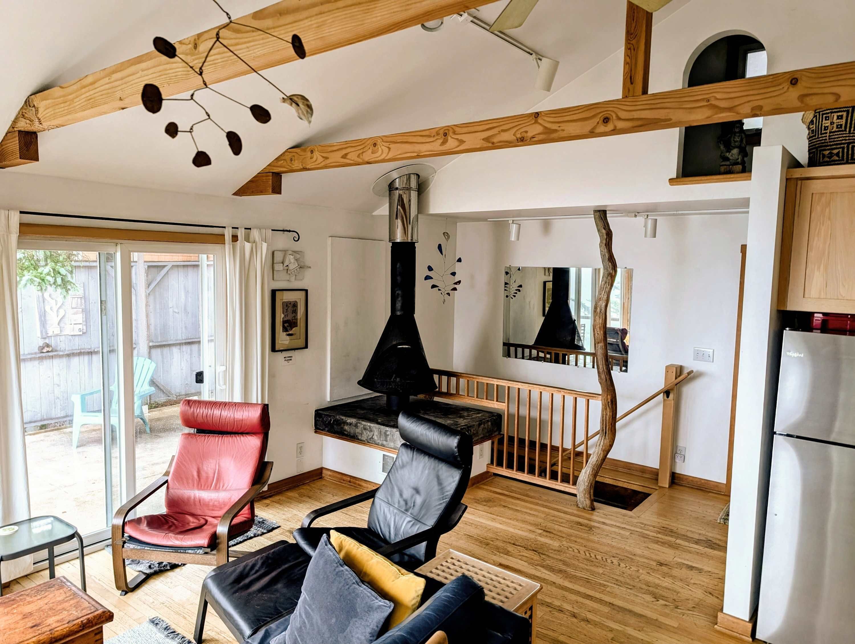 A modern living room with a fireplace, wooden beams, a red and black leather chair, and a kitchen area with a refrigerator.