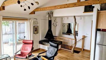 A modern living room with a fireplace, wooden beams, a red and black leather chair, and a kitchen area with a refrigerator.
