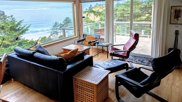 Ett mysigt vardagsrum med utsikt över havet, inrett med en svart lädersoffa, ett träbord och en röd fåtölj.