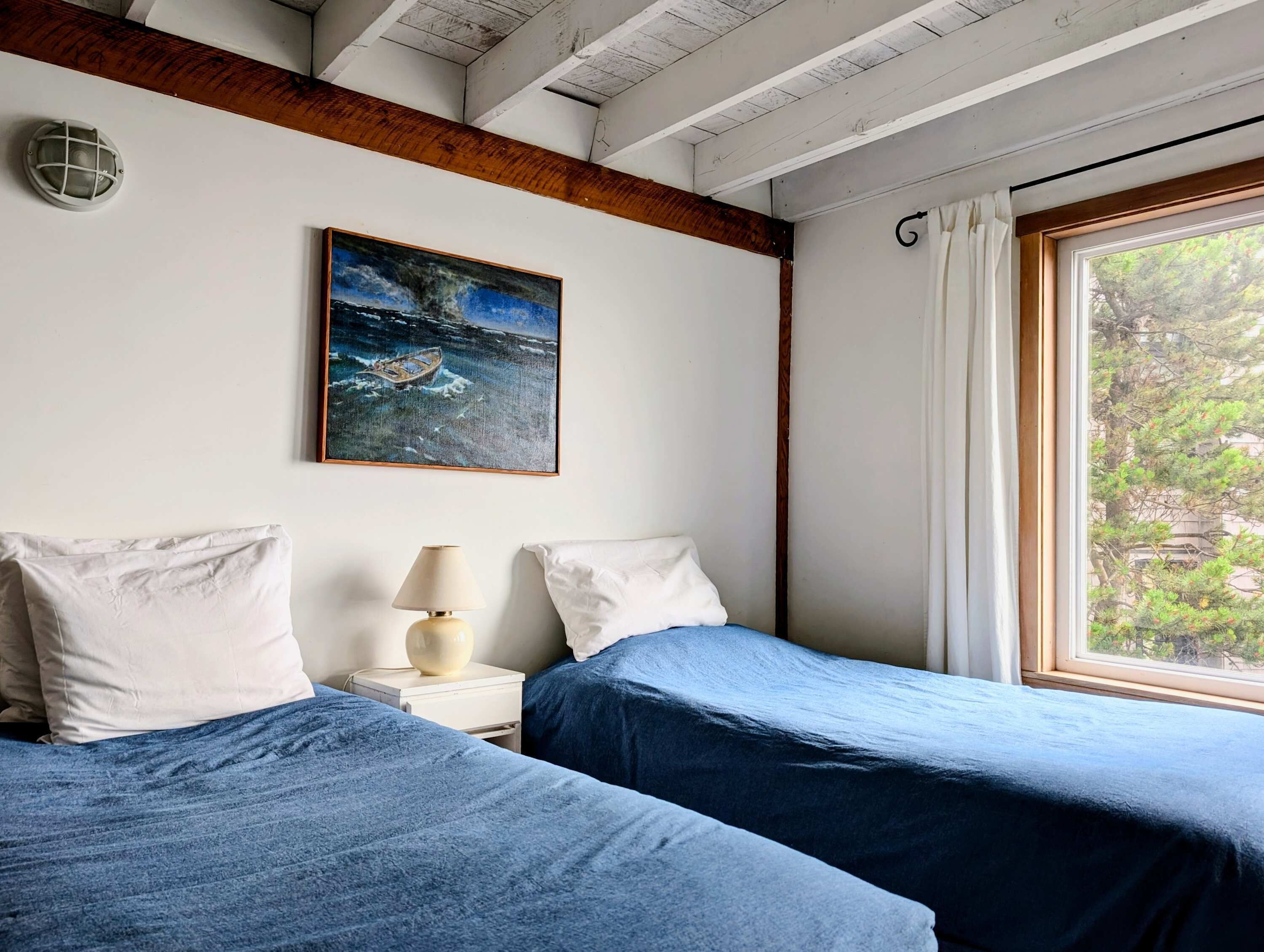 A room with two beds, a painting of a boat on water, a lamp, and a window with curtains.