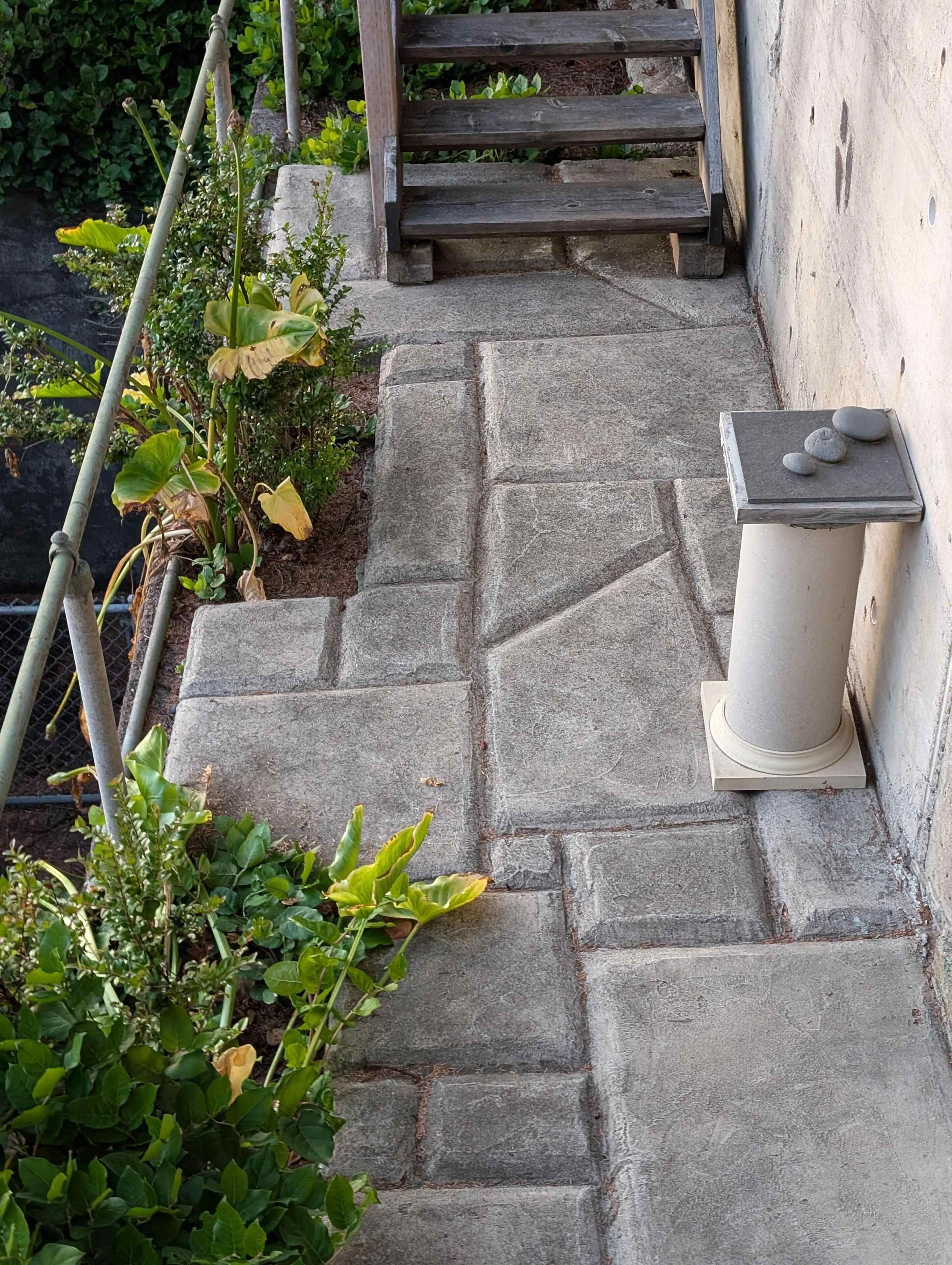 Una escalera de piedra que lleva a una terraza de madera, con un banco de piedra y una pequeña mesa con piedras.