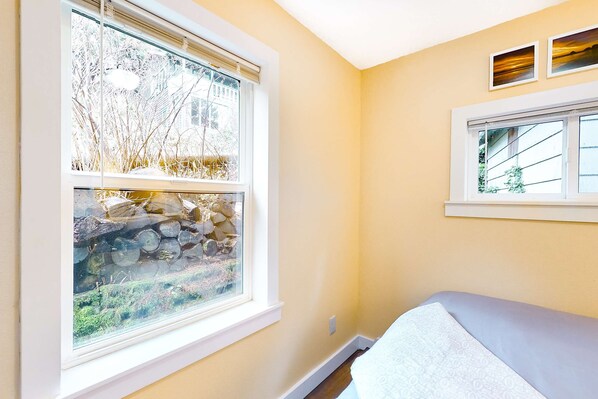 Un dormitorio con cama, ventana que ofrece vista a árboles y troncos, y pared con dos cuadros enmarcados.