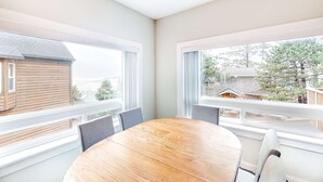 Un coin repas avec une table ronde en bois et quatre chaises, de grandes fenêtres à cadres blancs et une vue sur les maisons voisines.