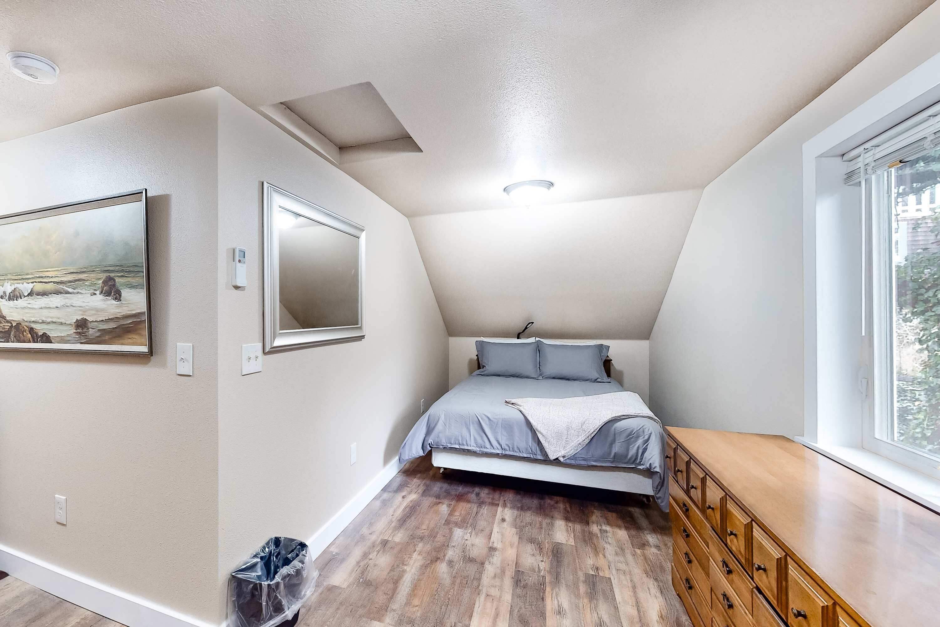A bedroom with a sloped ceiling, a bed with grey bedding, a wooden dresser, and a painting on the wall.