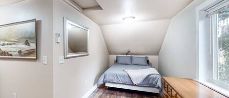 A bedroom with a sloped ceiling, a bed with grey bedding, a wooden dresser, and a painting on the wall.
