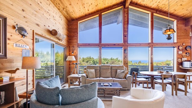 A cozy cabin interior with a large window, a sofa, a dining table, and a lamp. - Dog Friendly house with ocean view private hot tub wall of windows (Oceanside)