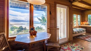 A cozy cabin interior with a wooden table and chairs, large windows with a view of the outdoors, and a comfortable seating area with a cushioned bench. - Hand Built Gem with Two Large Decks and Incredible Views 1 4 Mile to the Beach (Oceanside)