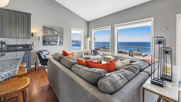 Een moderne woonkamer met een grijze modulaire bank, een keuken met barstoelen en grote ramen met uitzicht op zee.