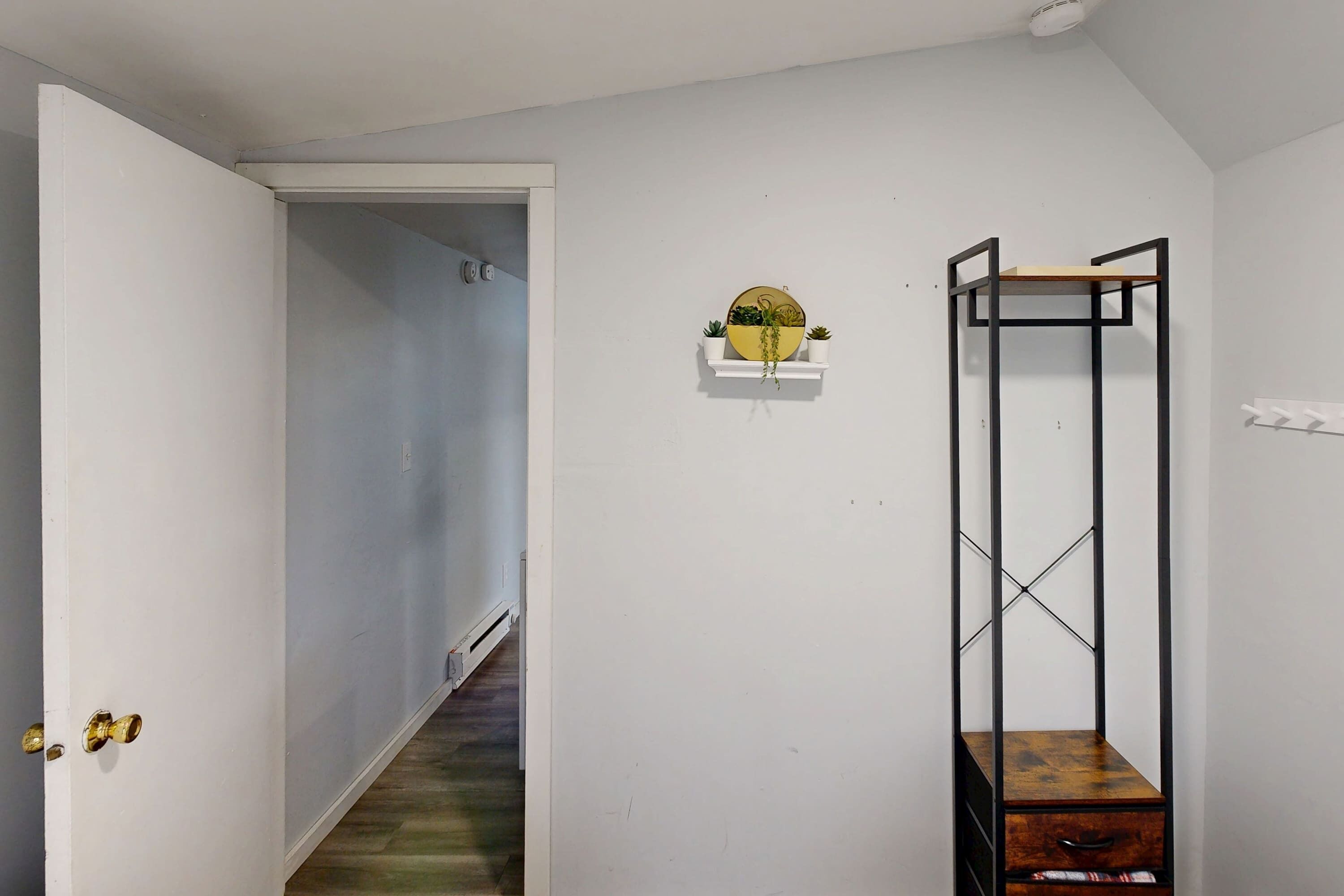 A hallway with a white door, a wooden staircase, a wall-mounted shelf with a round mirror, and a metal shelving unit with a wooden box.