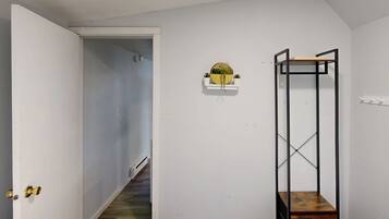 A hallway with a white door, a wooden staircase, a wall-mounted shelf with a round mirror, and a metal shelving unit with a wooden box.