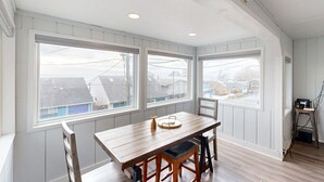 A room with a wooden dining table, two chairs, and large windows with a view of houses.