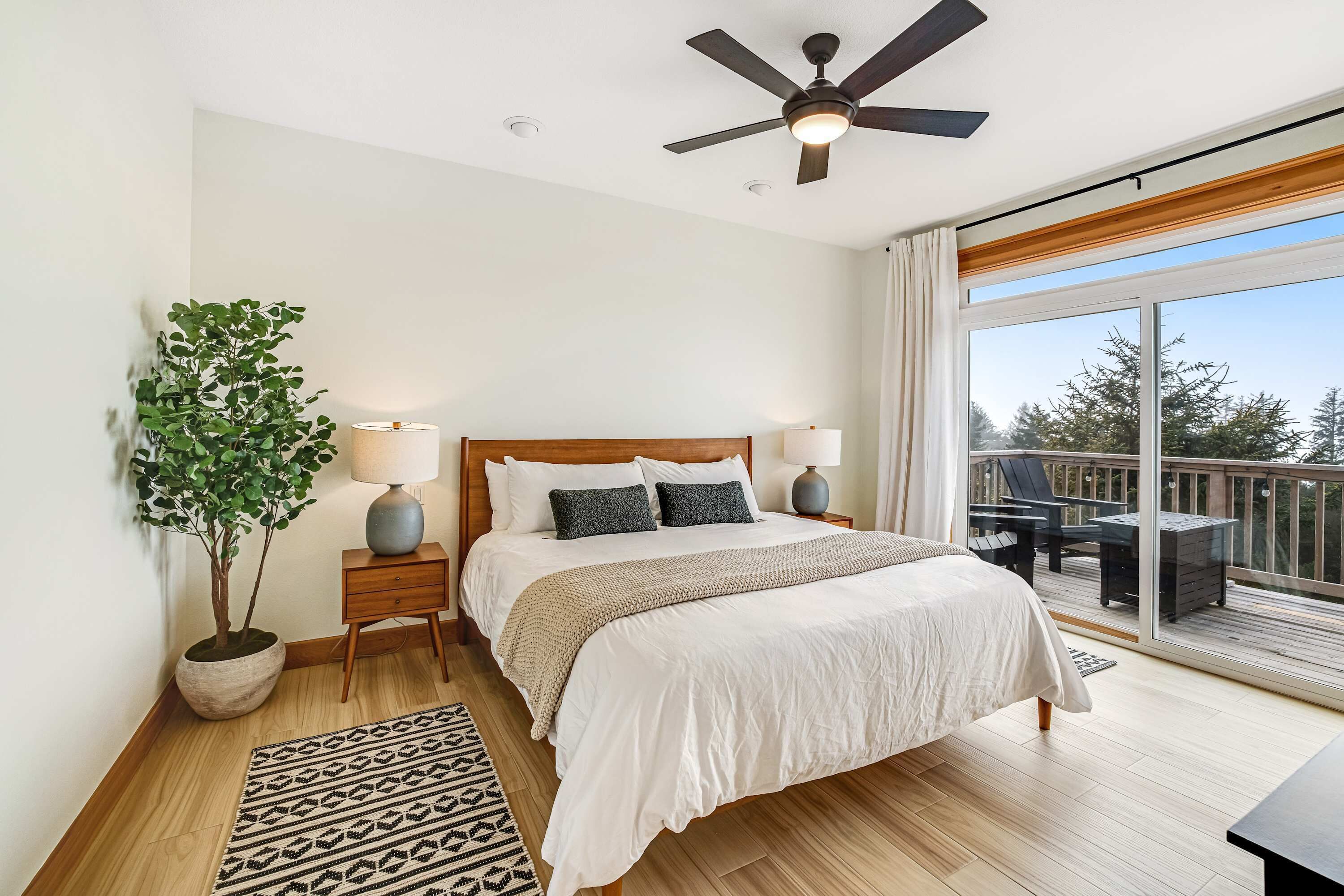 A bedroom with a bed, bedside lamps, a ceiling fan, a large window, and a balcony with outdoor furniture.