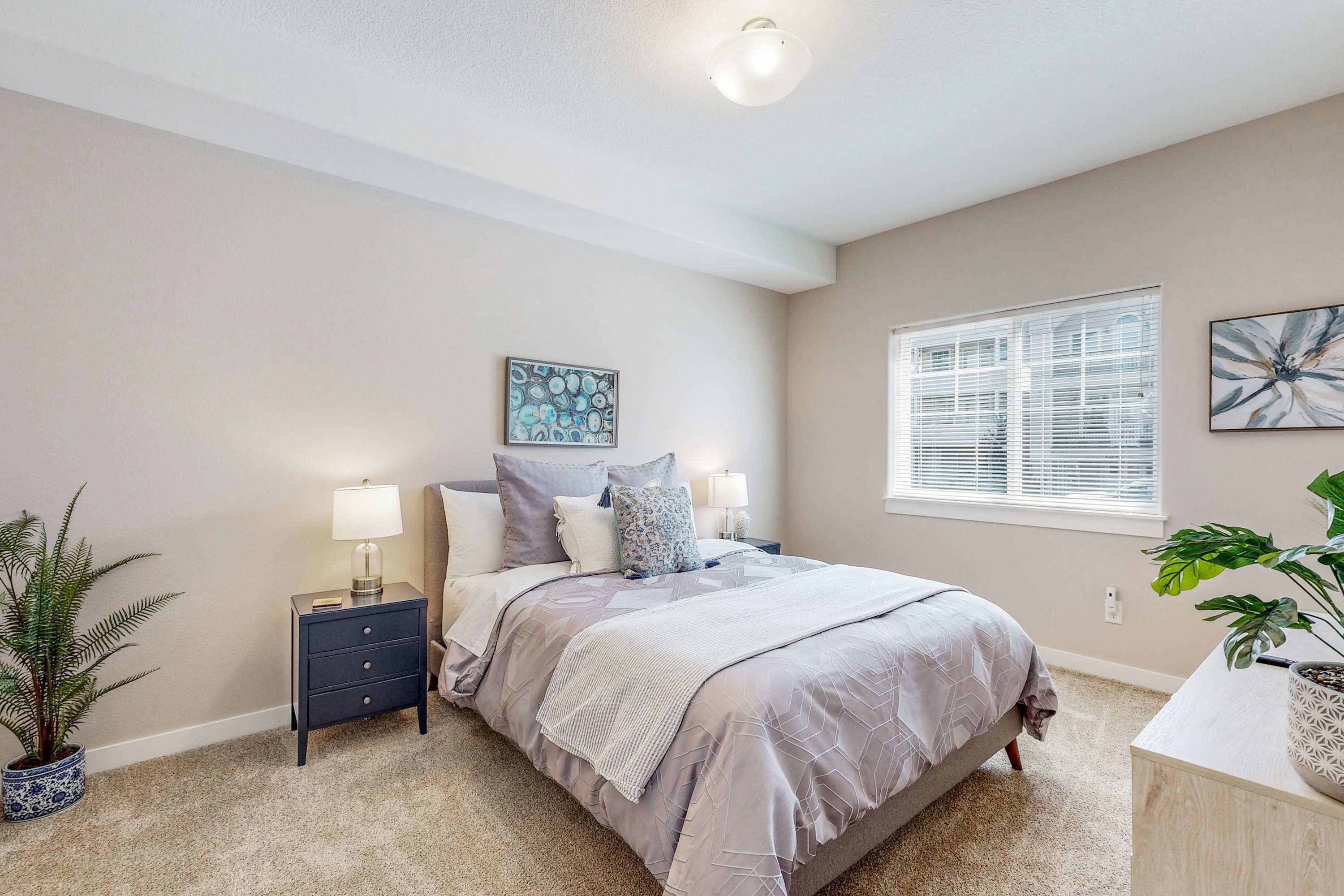 A bedroom with a bed, bedside tables, a nightstand, a potted plant, and a window with blinds.