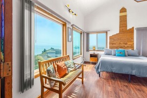 Een slaapkamer met een groot raam, een houten hoofdbord, een bed met kussens, een bankje en een nachtkastje.