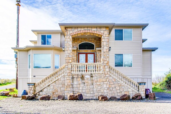 Une maison à deux étages avec une façade en pierre, une entrée centrale dotée d’un balcon et une allée bordée de rochers.