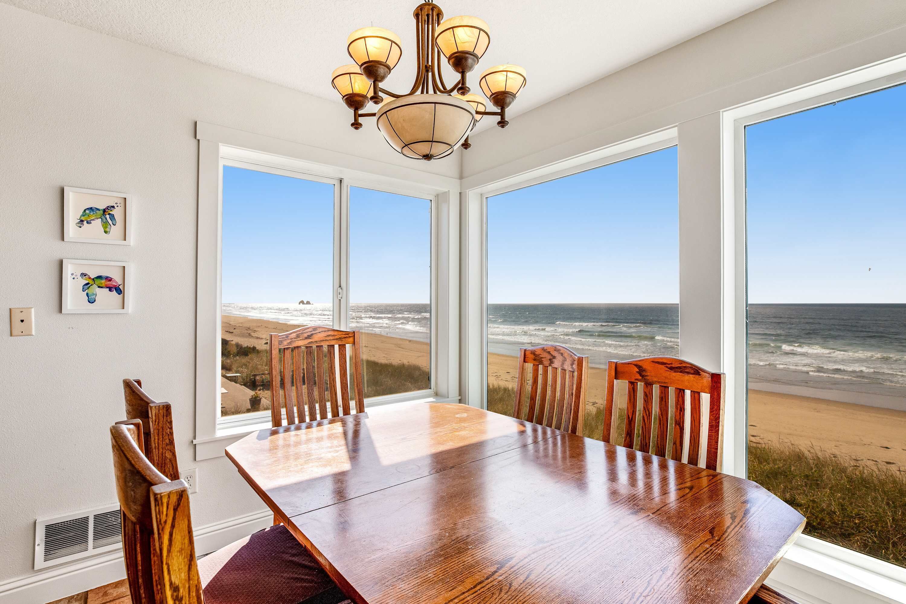 Un comedor con mesa y sillas de madera, ventanas amplias con vista a la playa y una araña.