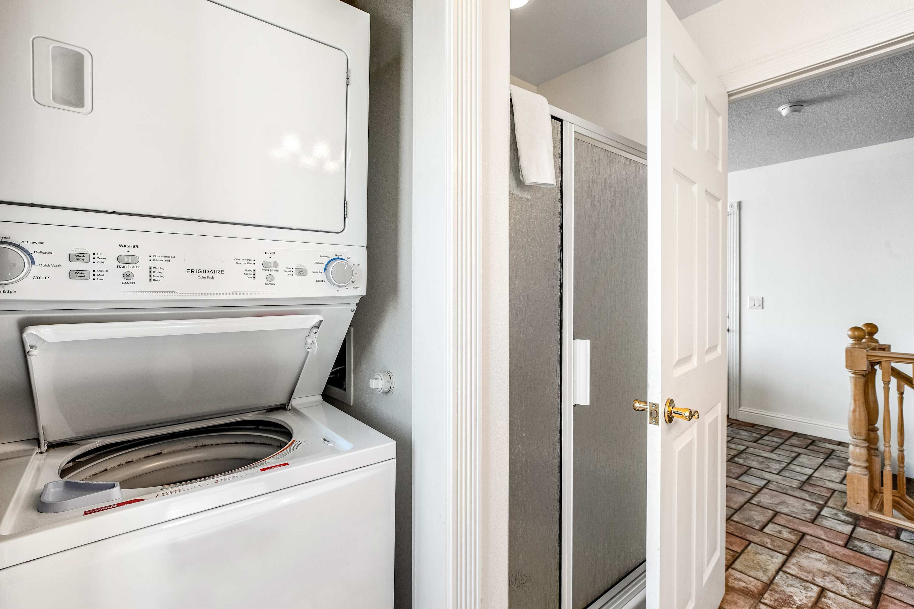 A laundry area with a front-loading washer and dryer, a door leading to a tiled hallway, and a wooden staircase.