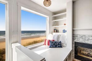 A room with a large window overlooking a beach, a fireplace, and a white shelf with books and decorative items. - Waterfront dog friendly home a few steps from the beach with hot tub (Rockaway Beach)