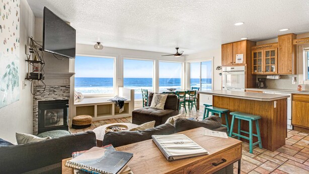 A spacious living area with a large window overlooking the ocean, a fireplace, and a kitchen with wooden cabinets and a bar area. - Charming dog friendly waterfront beach home with lovely patio (Rockaway Beach)